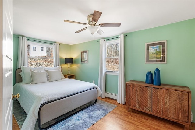 bedroom featuring a baseboard heating unit, multiple windows, wood finished floors, and a ceiling fan