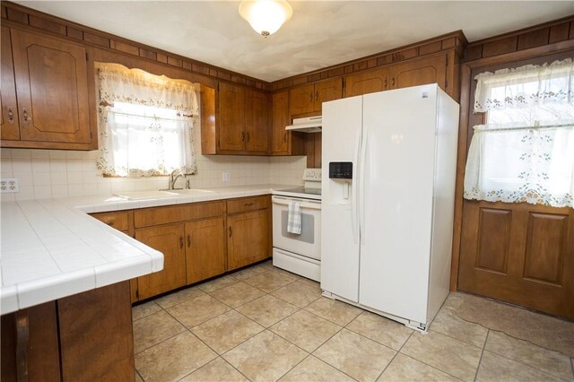 kitchen with white appliances, brown cabinetry, a sink, tile counters, and under cabinet range hood