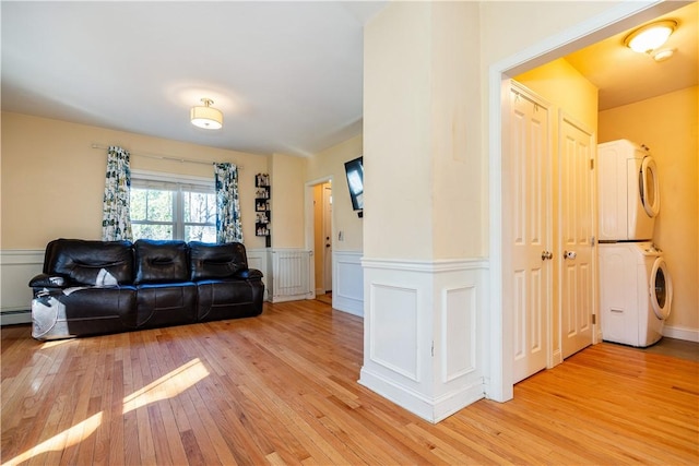 living area with stacked washing maching and dryer, wainscoting, light wood-style flooring, a decorative wall, and a baseboard heating unit