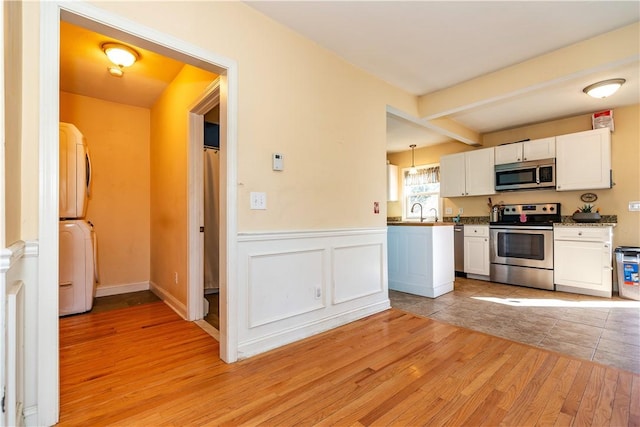 kitchen with a wainscoted wall, white cabinets, stacked washer / drying machine, appliances with stainless steel finishes, and light wood-type flooring