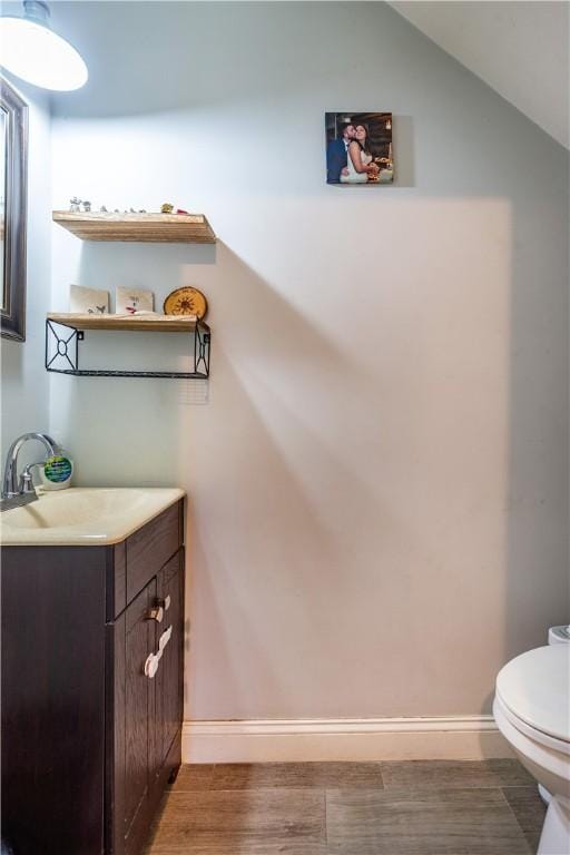 half bathroom featuring toilet, vanity, wood finished floors, lofted ceiling, and baseboards