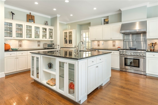kitchen featuring wood finished floors, a center island with sink, a sink, high end stove, and wall chimney range hood