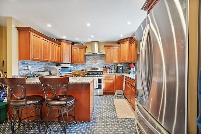 kitchen with wall chimney exhaust hood, stainless steel appliances, a peninsula, decorative backsplash, and brown cabinetry
