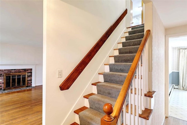 staircase featuring a brick fireplace, baseboard heating, baseboards, and wood finished floors