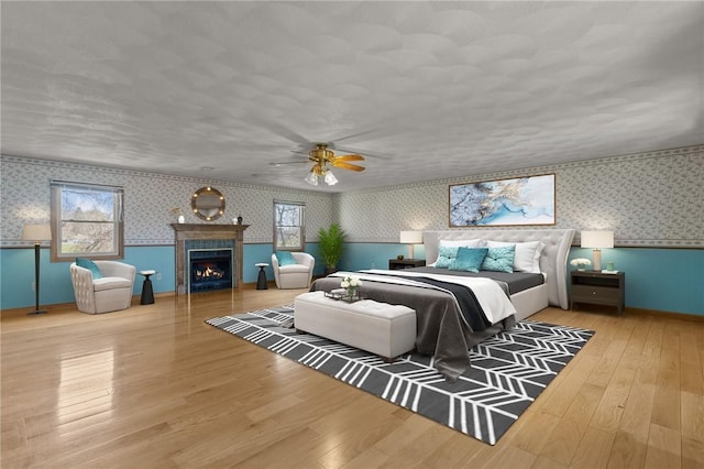 bedroom featuring baseboards, ceiling fan, hardwood / wood-style flooring, wallpapered walls, and a lit fireplace