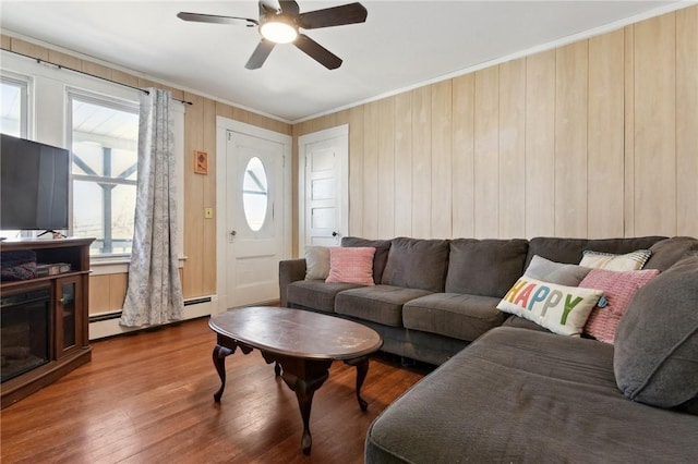 living area with wood finished floors, a baseboard heating unit, wooden walls, ceiling fan, and ornamental molding