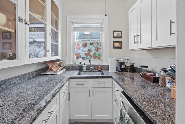 kitchen with dark stone counters, stainless steel dishwasher, glass insert cabinets, white cabinetry, and a sink