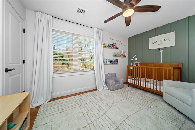 bedroom featuring ceiling fan, visible vents, a nursery area, wood finished floors, and baseboards