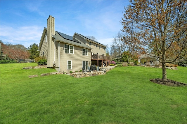 rear view of property with a chimney, a lawn, a wooden deck, roof mounted solar panels, and a patio