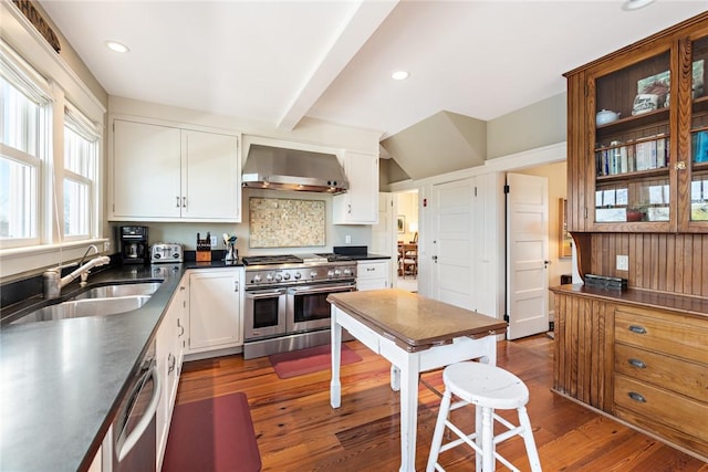 kitchen with appliances with stainless steel finishes, wall chimney range hood, dark countertops, a sink, and white cabinetry