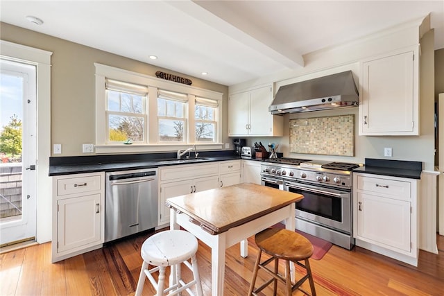 kitchen featuring appliances with stainless steel finishes, dark countertops, light wood finished floors, a sink, and wall chimney exhaust hood