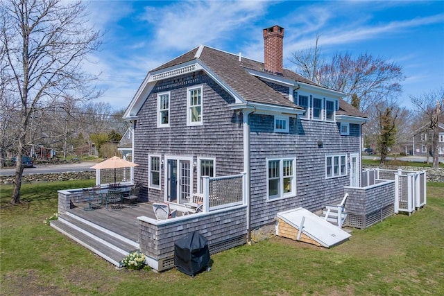 back of property with a chimney, a yard, a shingled roof, a wooden deck, and outdoor dining area