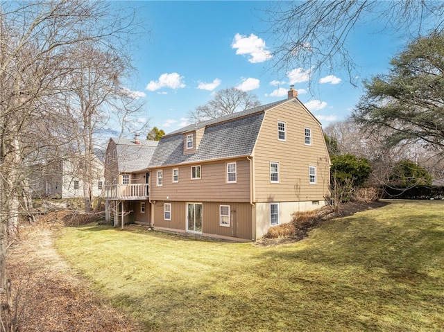 back of house featuring a gambrel roof, a chimney, a shingled roof, a yard, and a wooden deck