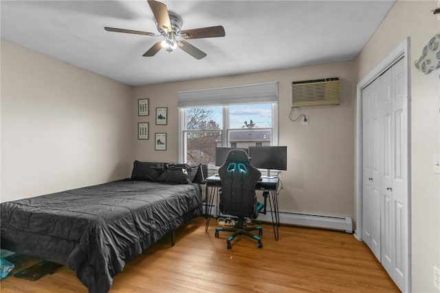 bedroom with an AC wall unit, baseboard heating, wood finished floors, a closet, and ceiling fan