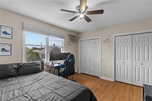 bedroom featuring light wood finished floors, baseboards, a wall mounted air conditioner, ceiling fan, and multiple closets
