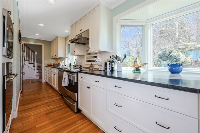 183 Twin Peninsula Ave, South Kingstown, RI 02879 - kitchen with under cabinet range hood, stainless steel appliances, ornamental molding, dark countertops, and light wood-type flooring