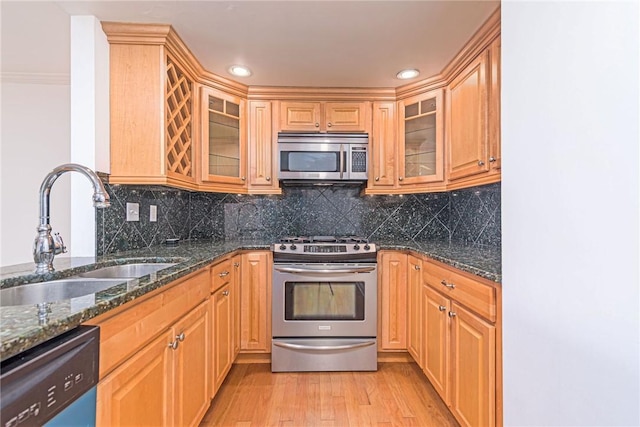 kitchen featuring light wood finished floors, stainless steel appliances, a sink, dark stone counters, and glass insert cabinets