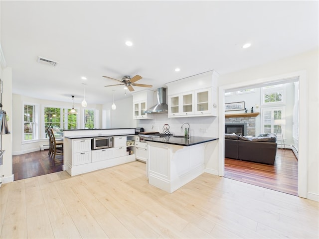 kitchen with a peninsula, stainless steel microwave, wall chimney exhaust hood, white cabinets, and dark countertops