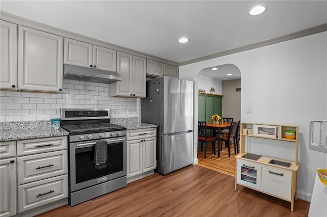 kitchen featuring light wood finished floors, stainless steel appliances, arched walkways, backsplash, and under cabinet range hood