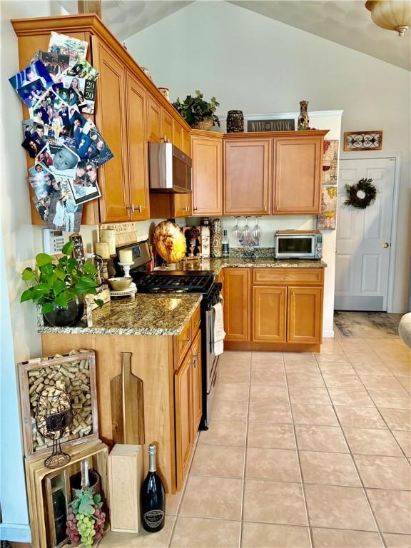 kitchen with vaulted ceiling, light tile patterned flooring, stone counters, brown cabinetry, and appliances with stainless steel finishes