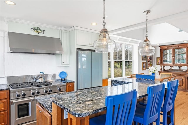 kitchen with recessed lighting, freestanding refrigerator, brown cabinetry, range with two ovens, and wall chimney exhaust hood