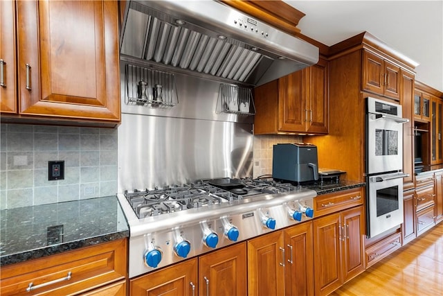 kitchen with stainless steel appliances, decorative backsplash, dark stone countertops, brown cabinetry, and wall chimney range hood