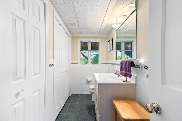 bathroom with tile patterned flooring, a drop ceiling, tile walls, toilet, and vanity