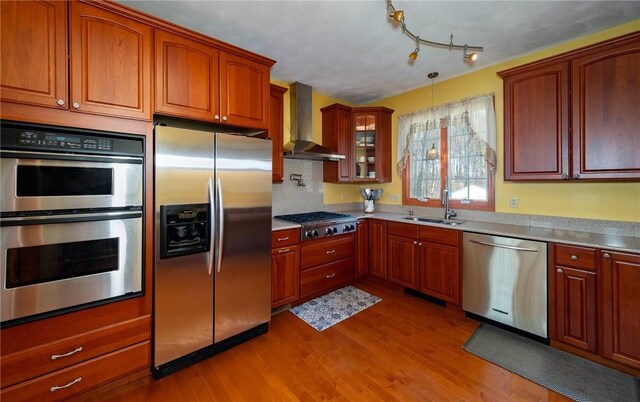kitchen featuring wood finished floors, glass insert cabinets, stainless steel appliances, wall chimney range hood, and a sink