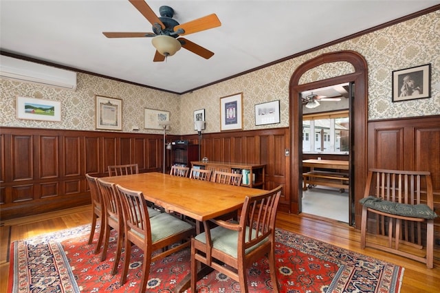dining room with a wall unit AC, a ceiling fan, ornamental molding, light wood finished floors, and wainscoting