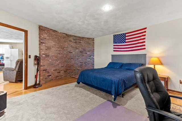 bedroom with a textured ceiling, brick wall, white refrigerator with ice dispenser, wood finished floors, and baseboards