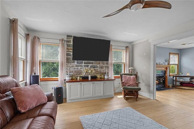 living room with a warm lit fireplace, ceiling fan, a wealth of natural light, and light wood-style floors