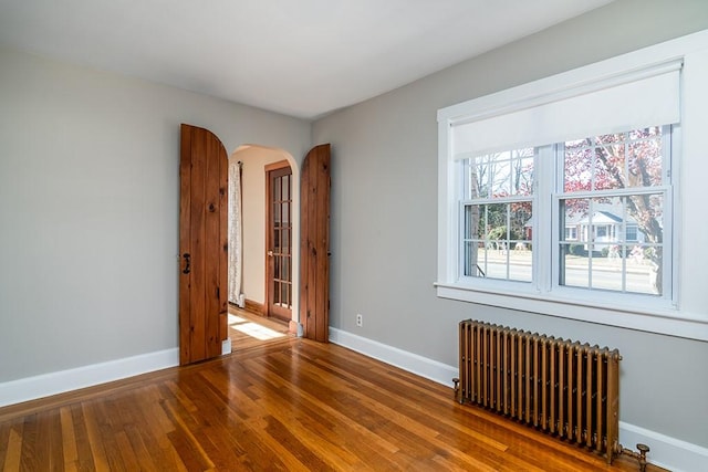 empty room featuring arched walkways, baseboards, wood finished floors, and radiator