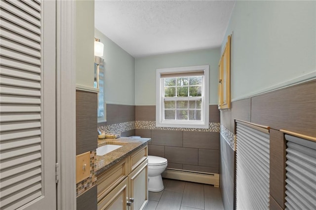 half bath featuring tile walls, tile patterned flooring, a wainscoted wall, a baseboard heating unit, and toilet