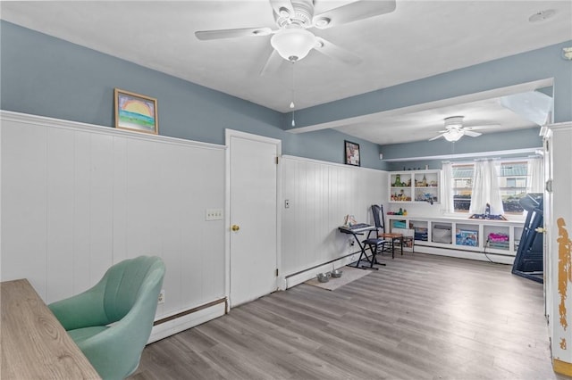 office area with ceiling fan, a baseboard heating unit, wood finished floors, and a wainscoted wall
