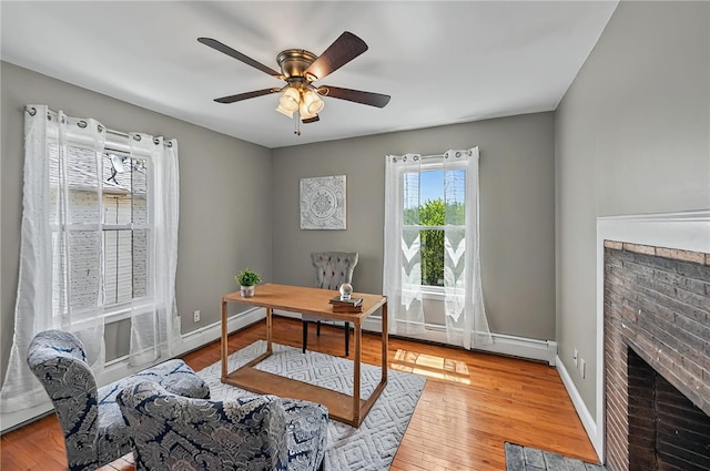 office area with a brick fireplace, a ceiling fan, baseboards, light wood-type flooring, and a baseboard heating unit
