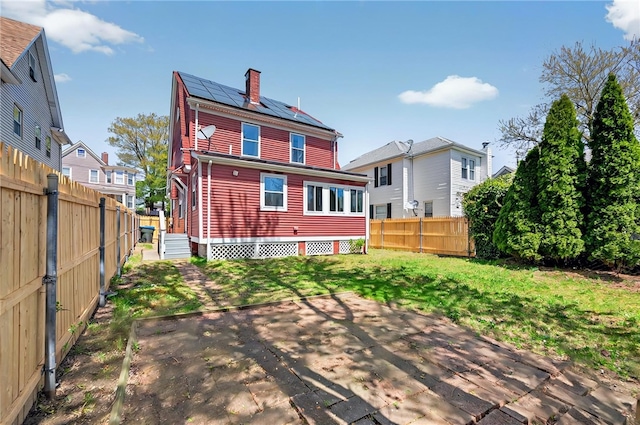 rear view of house with a patio, a fenced backyard, a chimney, roof mounted solar panels, and a yard