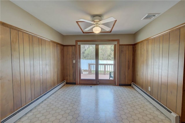 spare room featuring a ceiling fan, a baseboard heating unit, wood walls, and visible vents