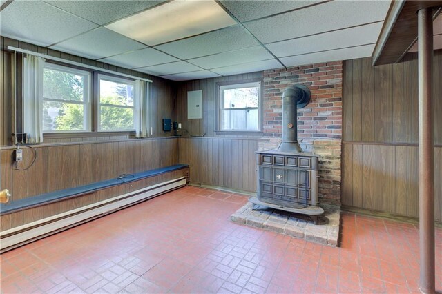 basement with wood walls, a wood stove, brick patterned floor, a drop ceiling, and baseboard heating