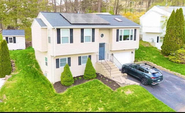 bi-level home featuring a garage, a front lawn, aphalt driveway, roof mounted solar panels, and a storage shed