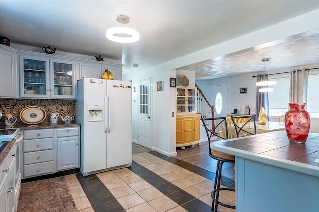 kitchen featuring dark tile patterned floors, decorative backsplash, white refrigerator with ice dispenser, glass insert cabinets, and tile counters