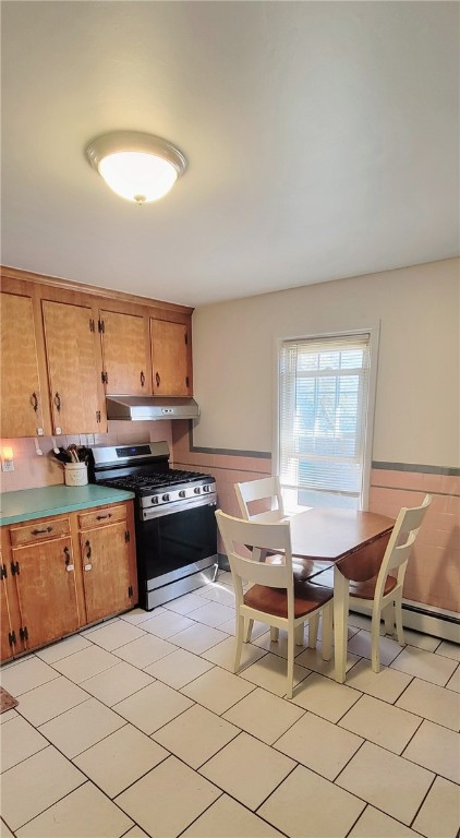 kitchen with under cabinet range hood, brown cabinets, wainscoting, and stainless steel gas stove