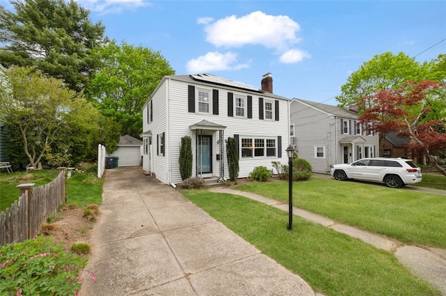 colonial-style house featuring fence, a front yard, roof mounted solar panels, an outbuilding, and a chimney