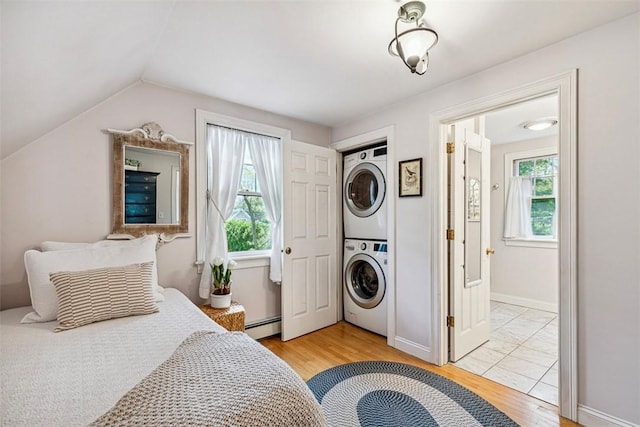 bedroom with a baseboard heating unit, stacked washing maching and dryer, lofted ceiling, and light wood-style floors