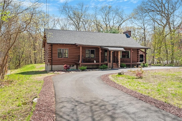 log-style house featuring roof with shingles, log siding, a porch, a chimney, and driveway