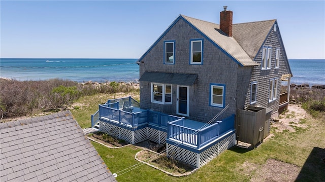 back of property featuring a chimney, a deck with water view, a lawn, a view of the beach, and a shingled roof