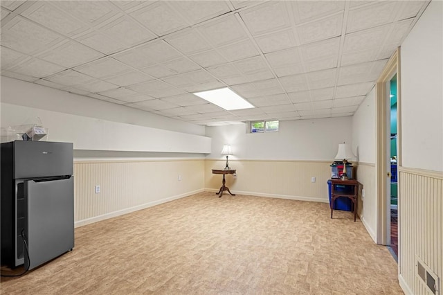 below grade area featuring baseboards, freestanding refrigerator, a drop ceiling, and a wainscoted wall