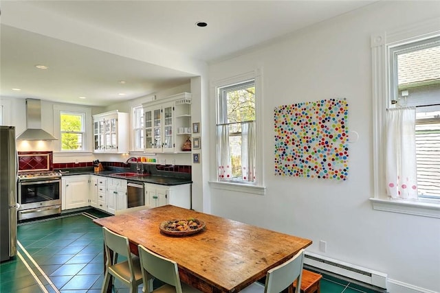 kitchen with wall chimney range hood, appliances with stainless steel finishes, a sink, a baseboard heating unit, and a wealth of natural light