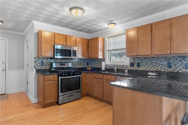 kitchen with appliances with stainless steel finishes, a sink, brown cabinetry, dark stone counters, and light wood-type flooring