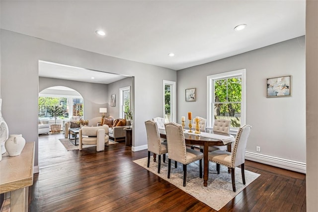 dining space featuring arched walkways, baseboard heating, dark wood-style flooring, and a wealth of natural light