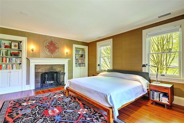bedroom featuring crown molding, a brick fireplace, baseboards, wood finished floors, and visible vents